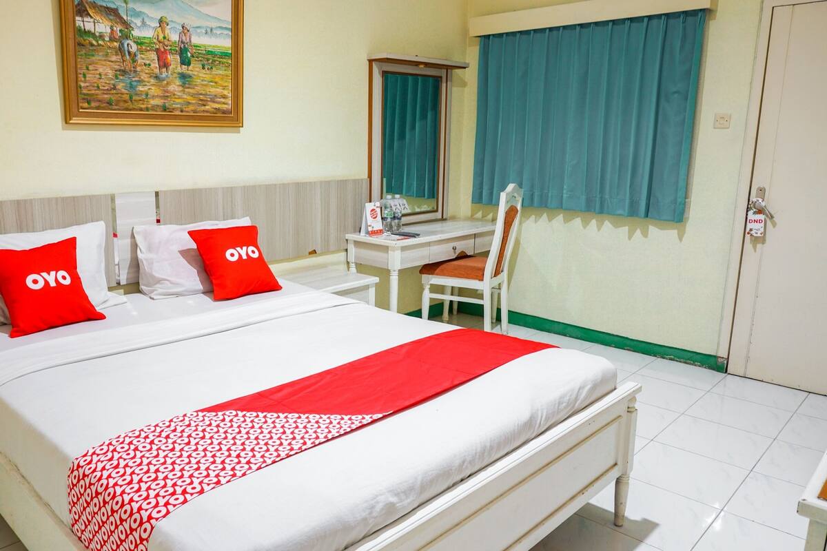 A hotel room with a bed, two nightstands, a desk, a chair, a window with curtains, and a painting on the wall.