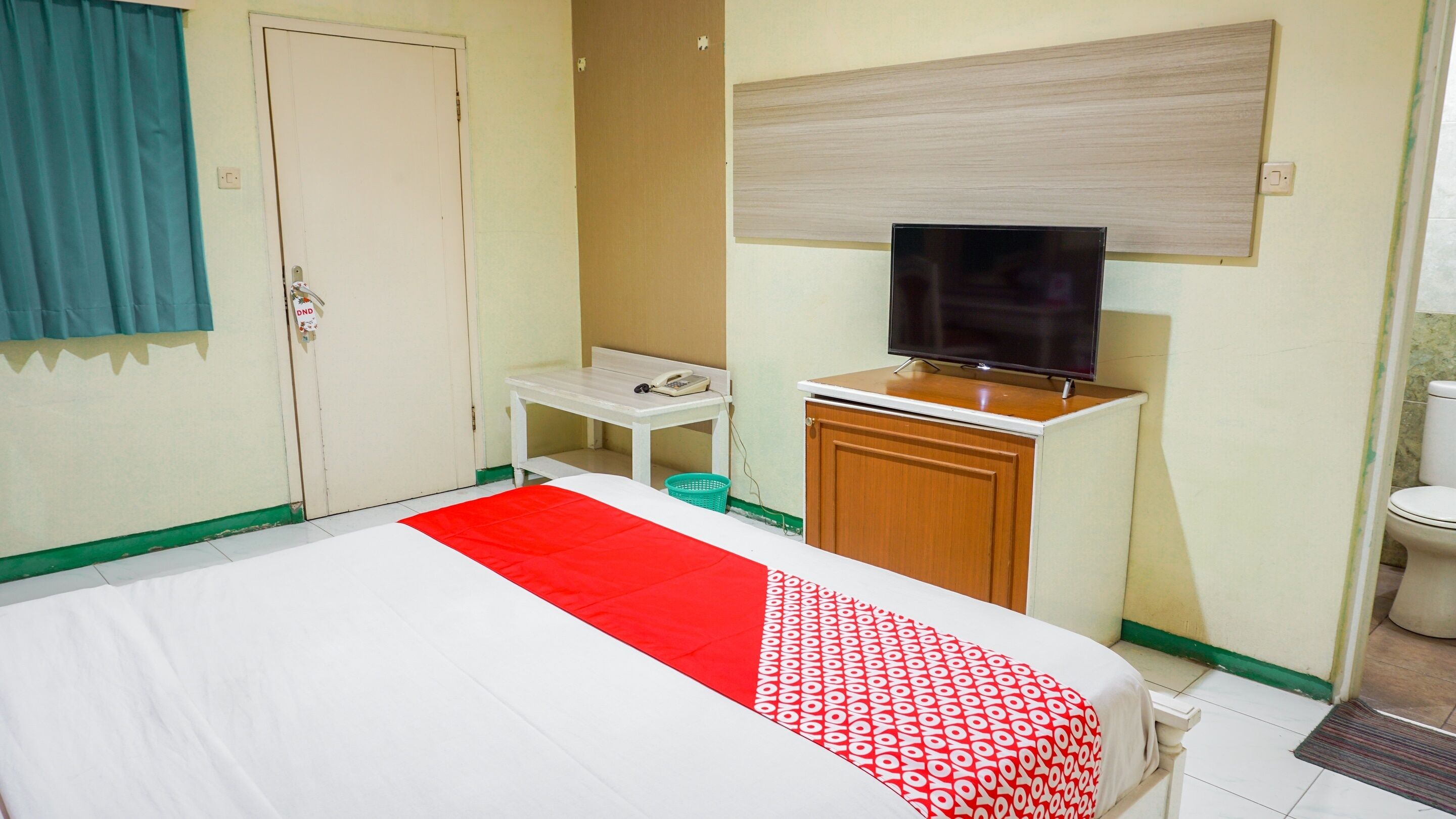 A hotel room with a bed, a small table with a phone, a television on a wooden cabinet, and a toilet.