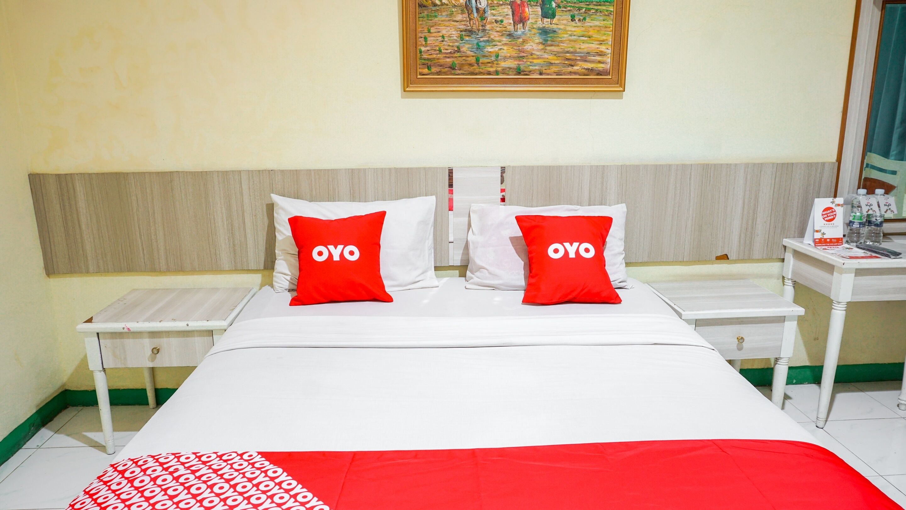 A hotel room with a bed, two nightstands, a painting on the wall, and a small table with a water bottle and a sign.