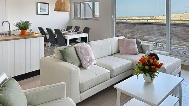 Een moderne woonkamer met een witte bank, een eethoek met stoelen, een keuken en grote ramen met uitzicht op het strand.