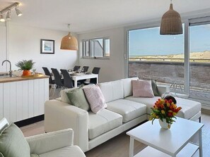 A modern living room with a white sofa, a dining area with chairs, a kitchen area, and large windows offering a view of the beach.