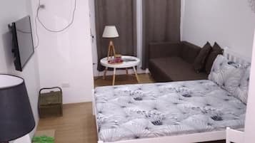 A compact bedroom with a single bed, a small table, a lamp, a TV, and a window with curtains.