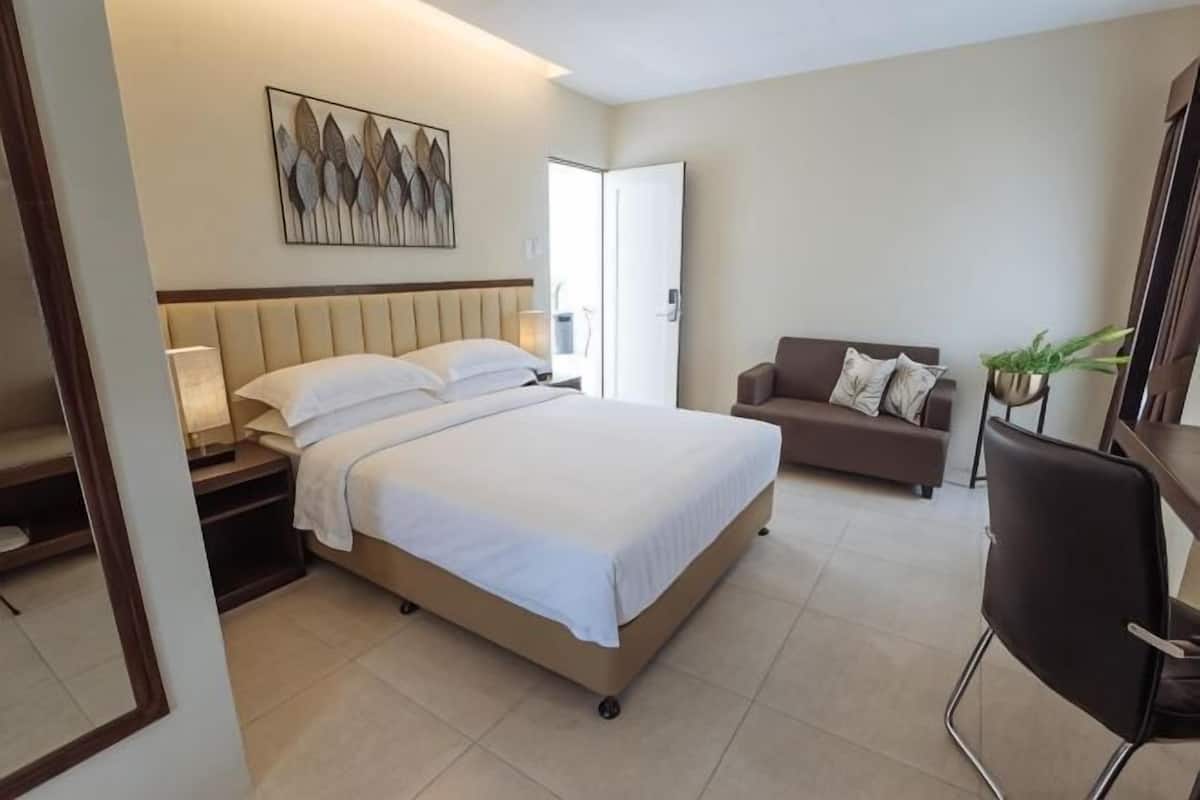 A hotel room with a bed, a sofa, a chair, a nightstand, a lamp, and a painting on the wall.