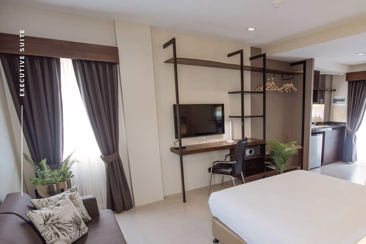 A hotel room with a bed, a desk with a TV, a chair, a sofa, and a small kitchenette.