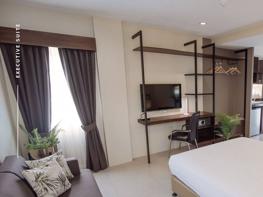 A hotel room with a bed, a desk with a TV, a chair, a sofa, and a small kitchenette.