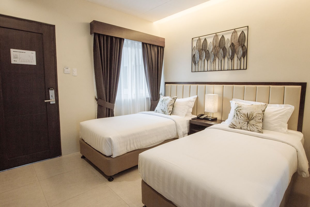 A hotel room with two beds, a nightstand, a lamp, a window with curtains, a door, and a wall-mounted artwork.