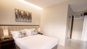 A neatly arranged bedroom with a bed, nightstands, a wall-mounted artwork, and a hallway with an air conditioning unit. - Rovira Suites (Dumaguete)