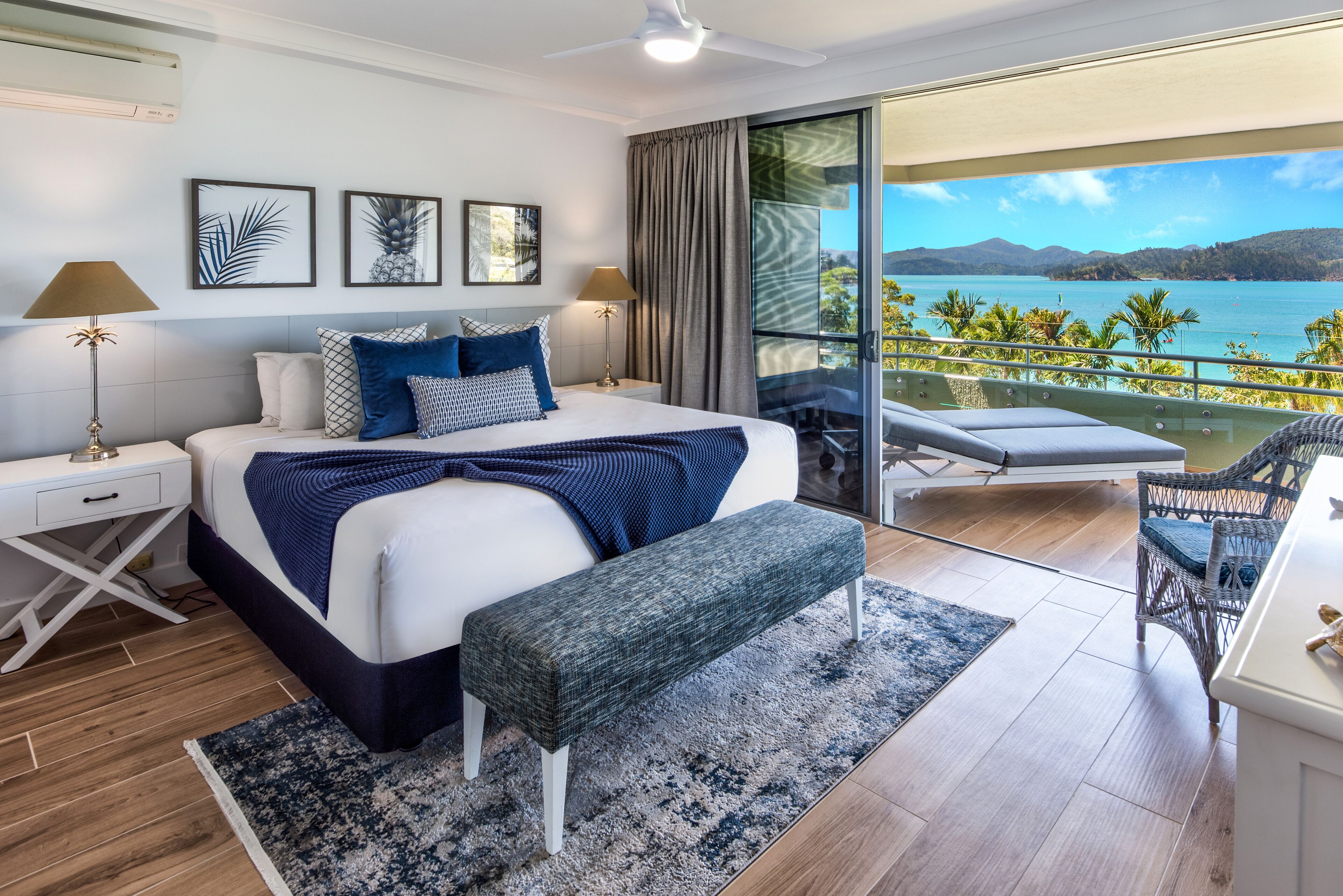 A modern bedroom with a large bed, bedside tables, a bench, and a balcony with a view of the sea and mountains.