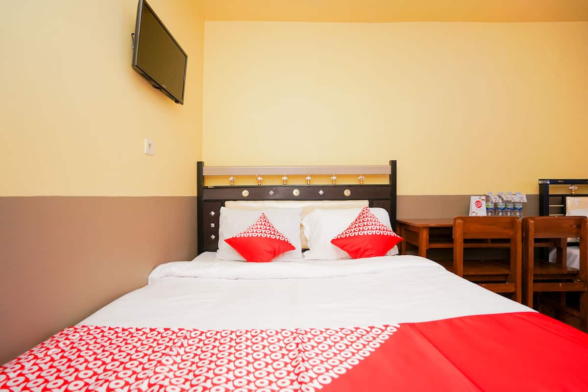 Ein Hotelzimmer mit einem Bett, zwei roten Kissen, einem an der Wand befestigten Fernseher und einem kleinen Tisch mit einer Flasche und einer Schachtel.