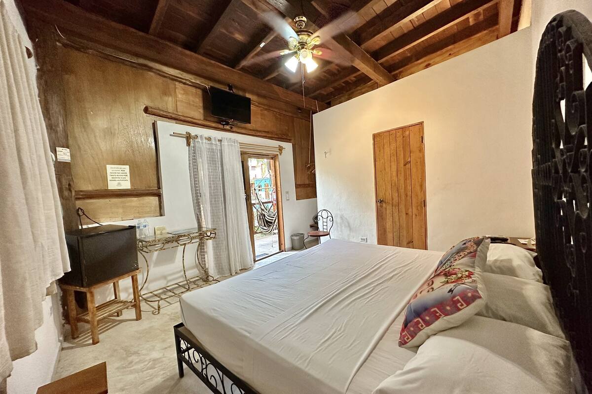 A bedroom with a bed, a desk, a chair, a ceiling fan, and a wooden door.