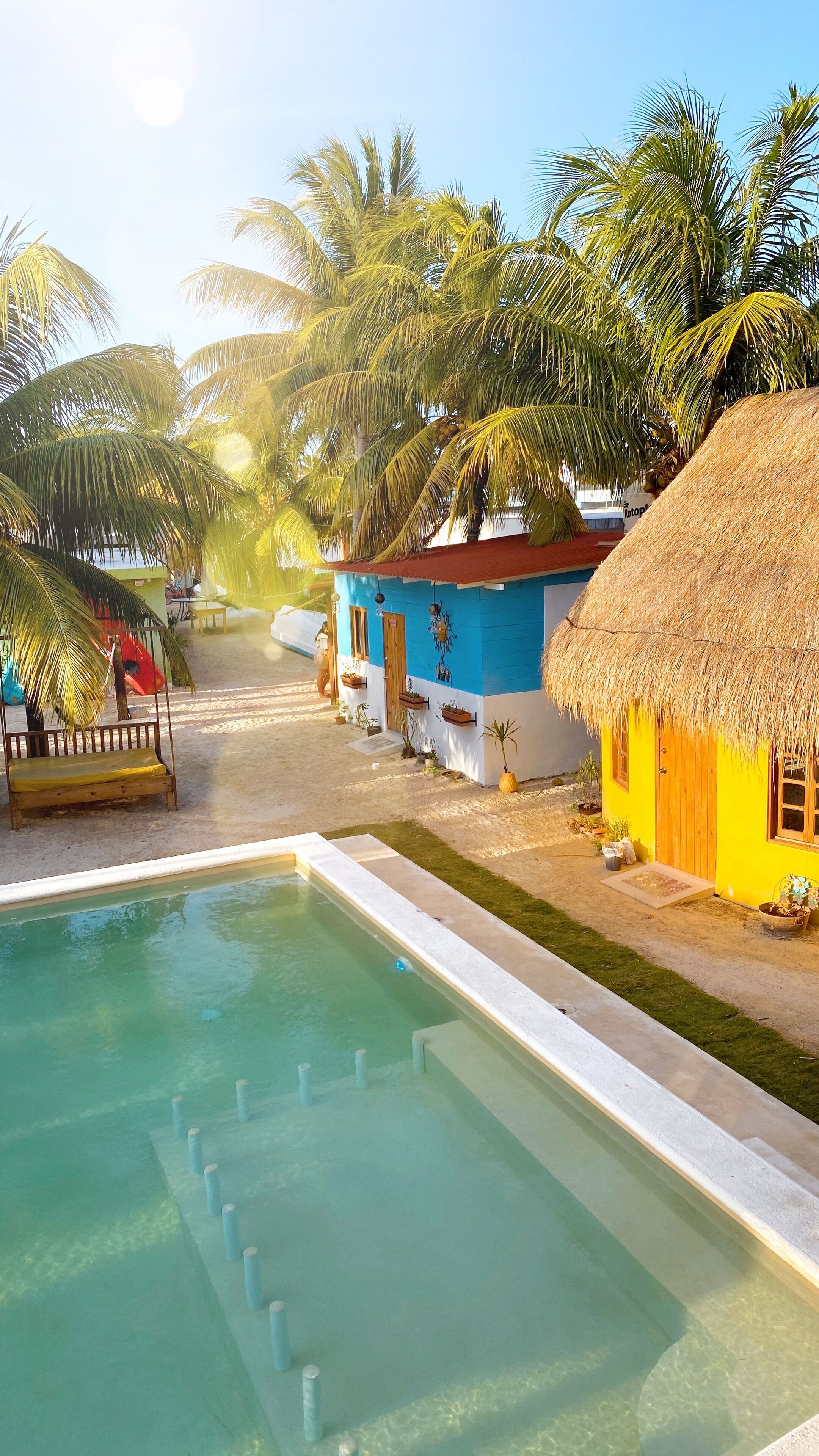 A pool area with a clear blue pool, surrounded by palm trees and thatched-roof buildings.