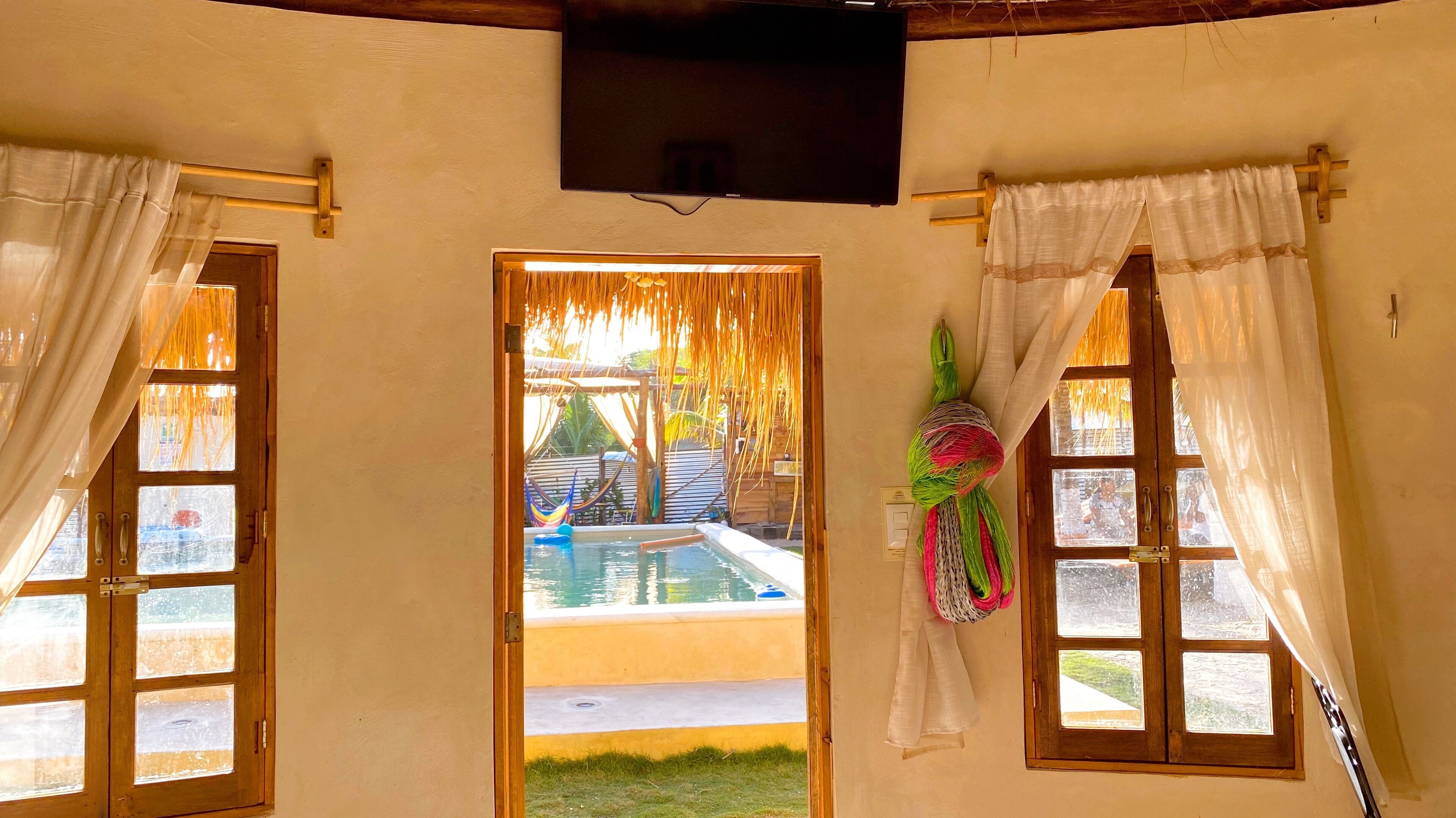 A room with a wooden door leading to a pool area, a television mounted on the wall, and a hammock hanging outside.