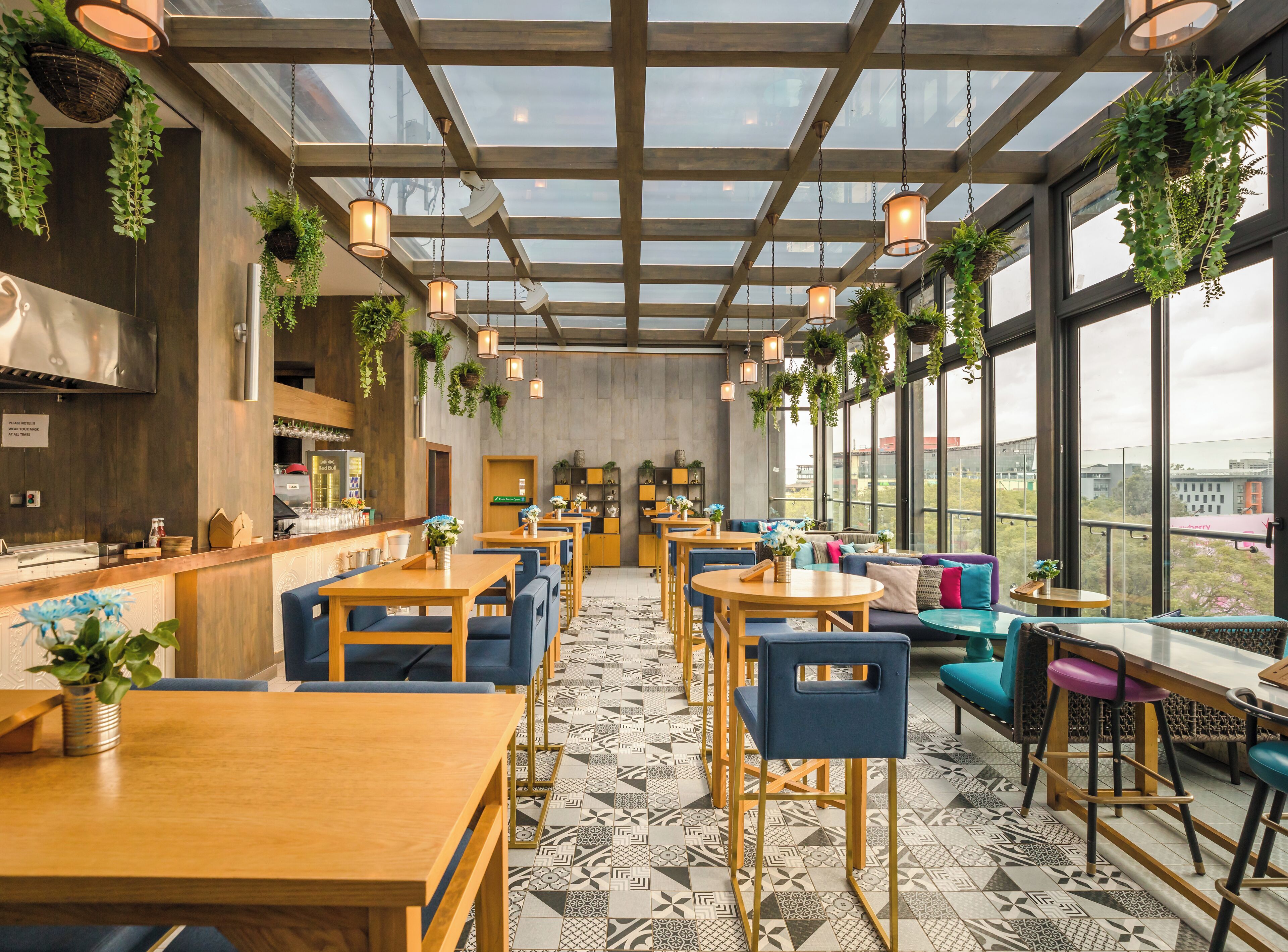 A modern restaurant with wooden tables and blue chairs, hanging plants, and large windows offering a city view.