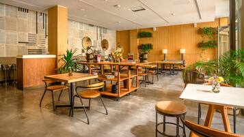 Un café moderne avec des tables et des chaises en bois, un comptoir où sont exposés des journaux et des plantes en pot.