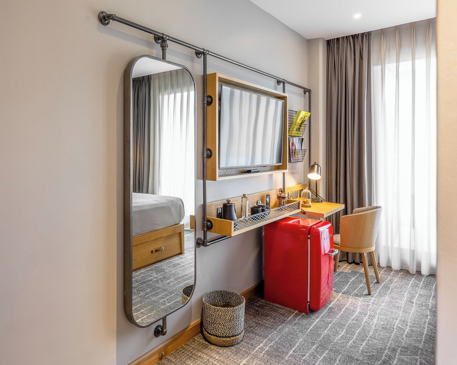 Una habitación de hotel con cama, un escritorio con silla, una maleta roja, un espejo y una ventana con cortinas.