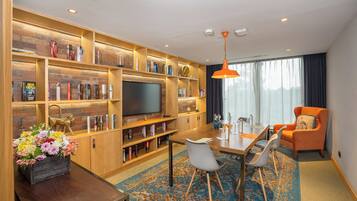 A modern hotel room with a wooden bookshelf, a flat-screen TV, a dining area with a table and chairs, and a large window with curtains.