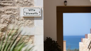 Een bord met de tekst 'Villa Stamatis' op een stenen muur, met uitzicht op zee door een open deur.