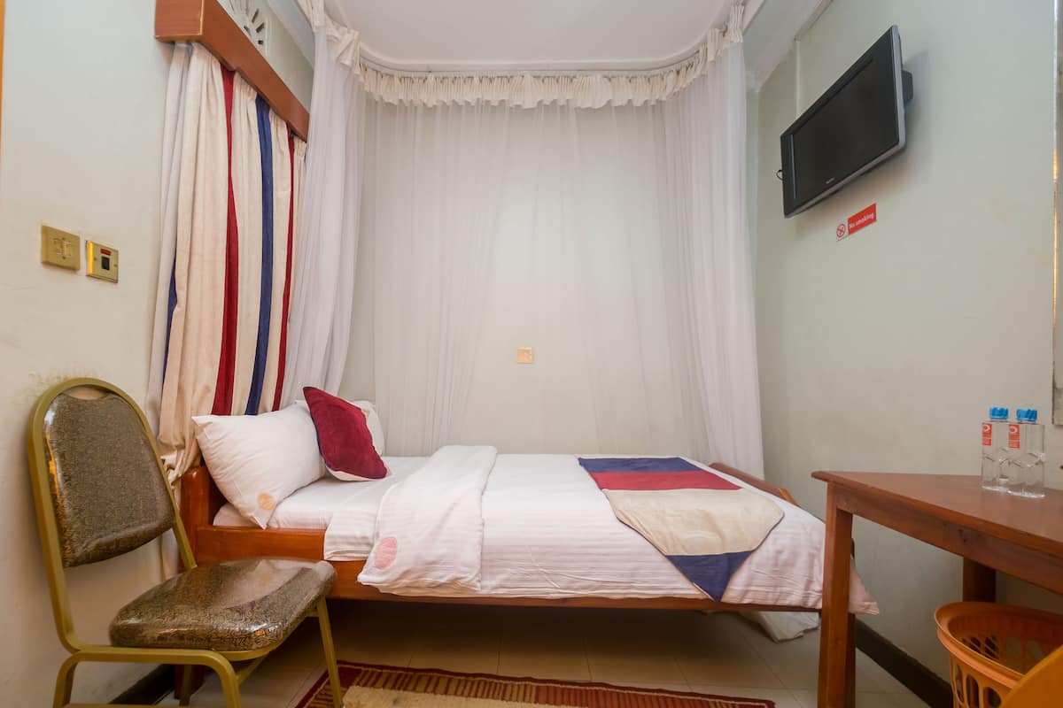 A small, neatly arranged bedroom with a single bed, a chair, a table, and a TV mounted on the wall.