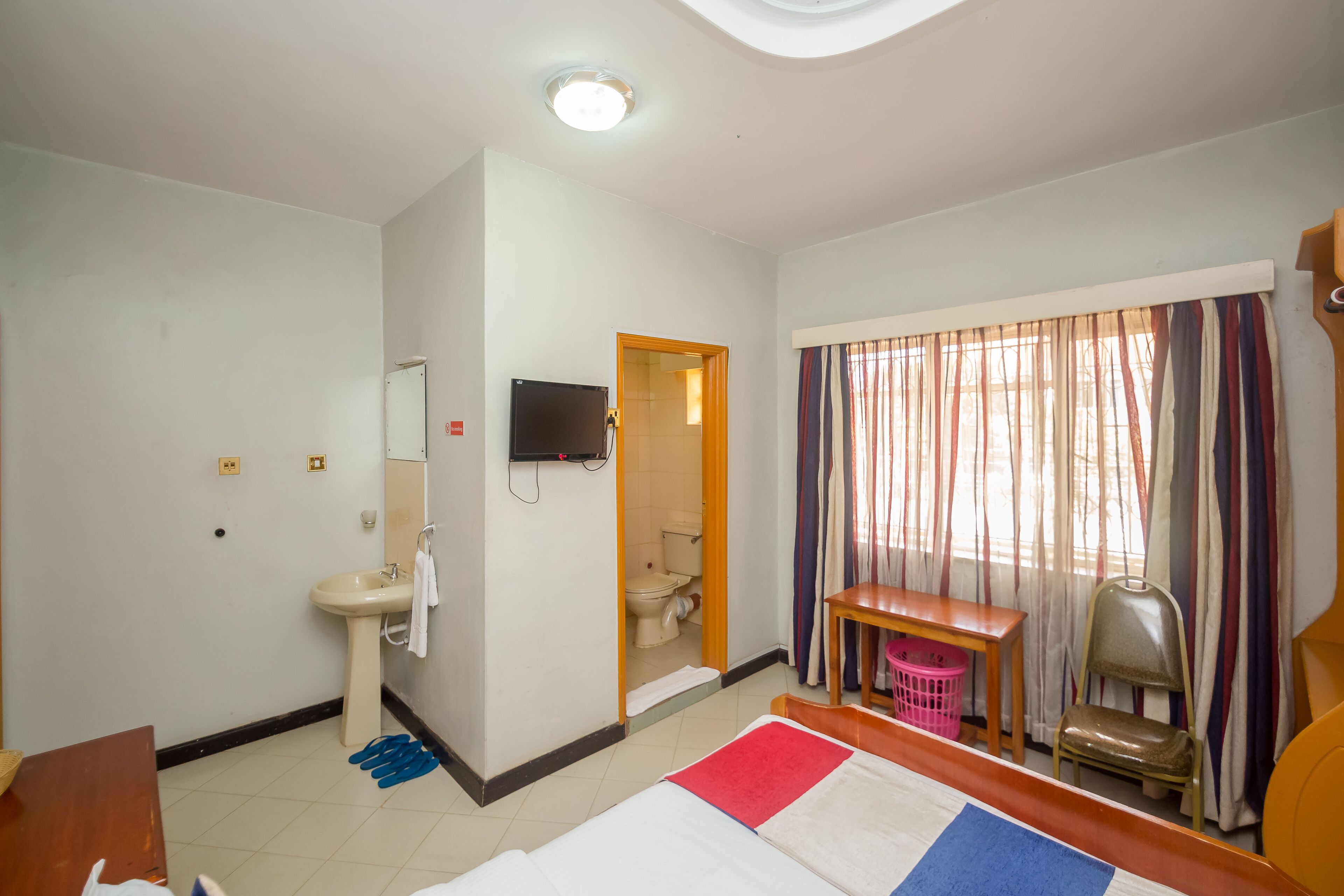Una habitación de hotel con cama, lavamanos, espejo, televisor, ventana con cortinas y una pequeña mesa.