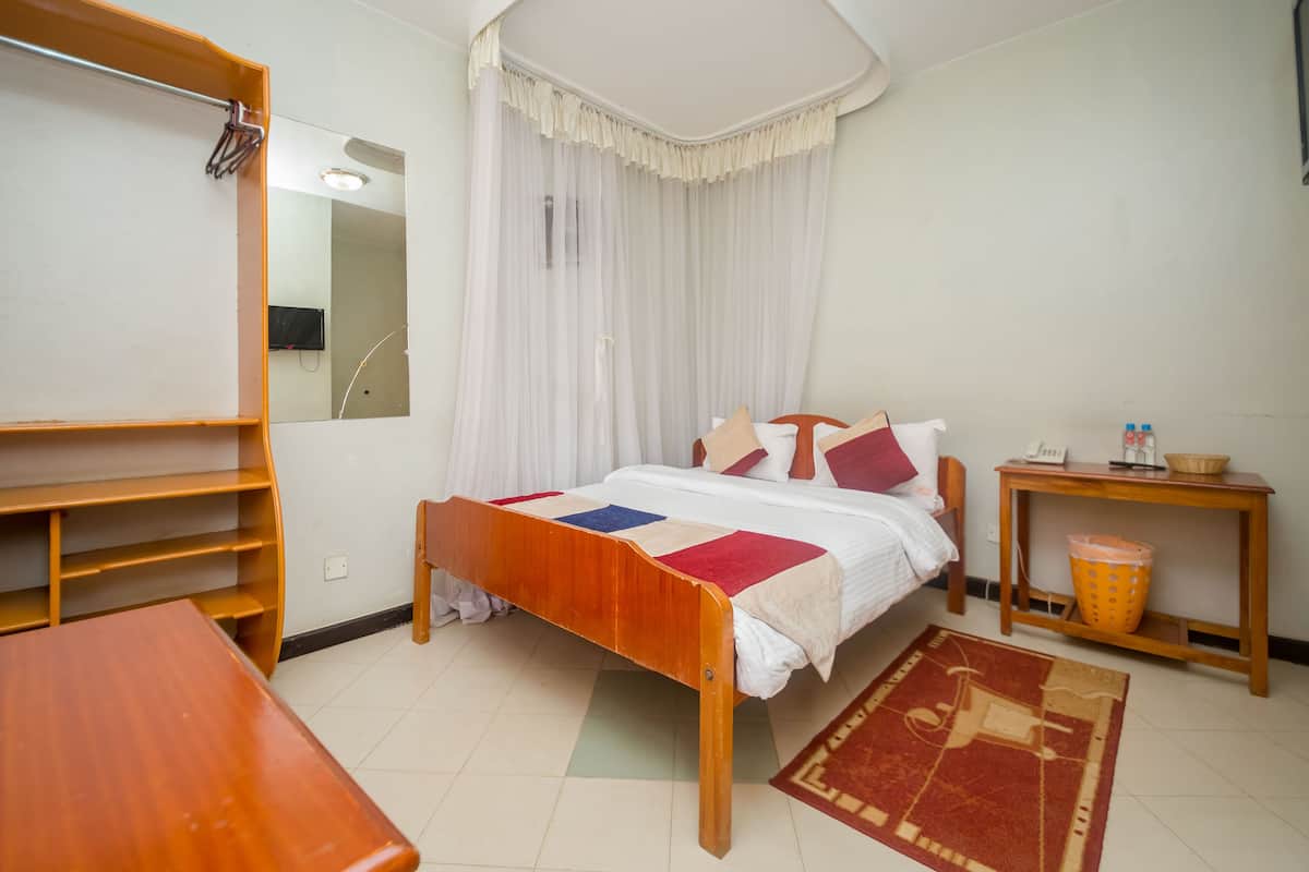 A bedroom with a wooden bed, a small table, a nightstand, a mirror, a wardrobe, and a window with sheer curtains.
