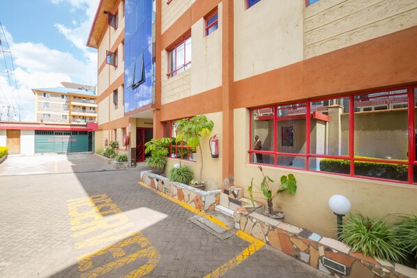 A multi-story building with a ground-level entrance, potted plants, and a clear pathway marked with yellow lines. - Jupiter Guest Resort Langata (Nairobi)