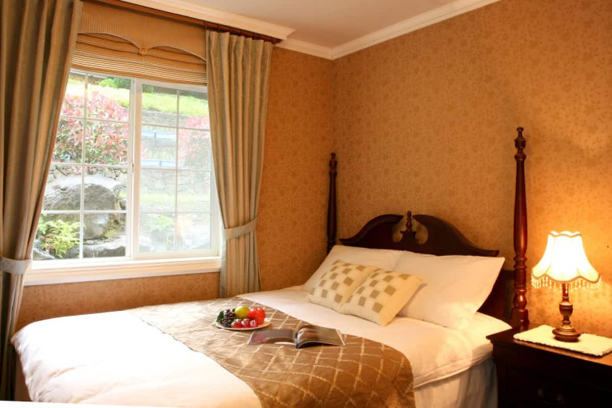 A bedroom with a bed, a nightstand with a lamp, a window with curtains, and a view of greenery outside.