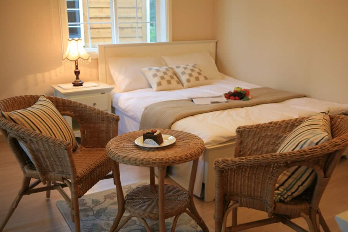 A neatly arranged bedroom with a bed, two wicker chairs, a small round table with a plate of food, and a bedside lamp.