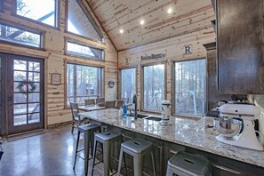 A spacious kitchen with a large island, wooden walls, and large windows offering views of the surrounding forest.