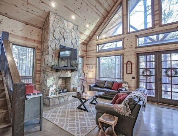 A spacious log cabin interior with a stone fireplace, a leather sofa, and large windows offering views of the surrounding forest.