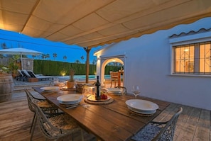 Un área de comedor al aire libre con una mesa de madera dispuesta para cuatro personas, rodeada de sillas, bajo un toldo.