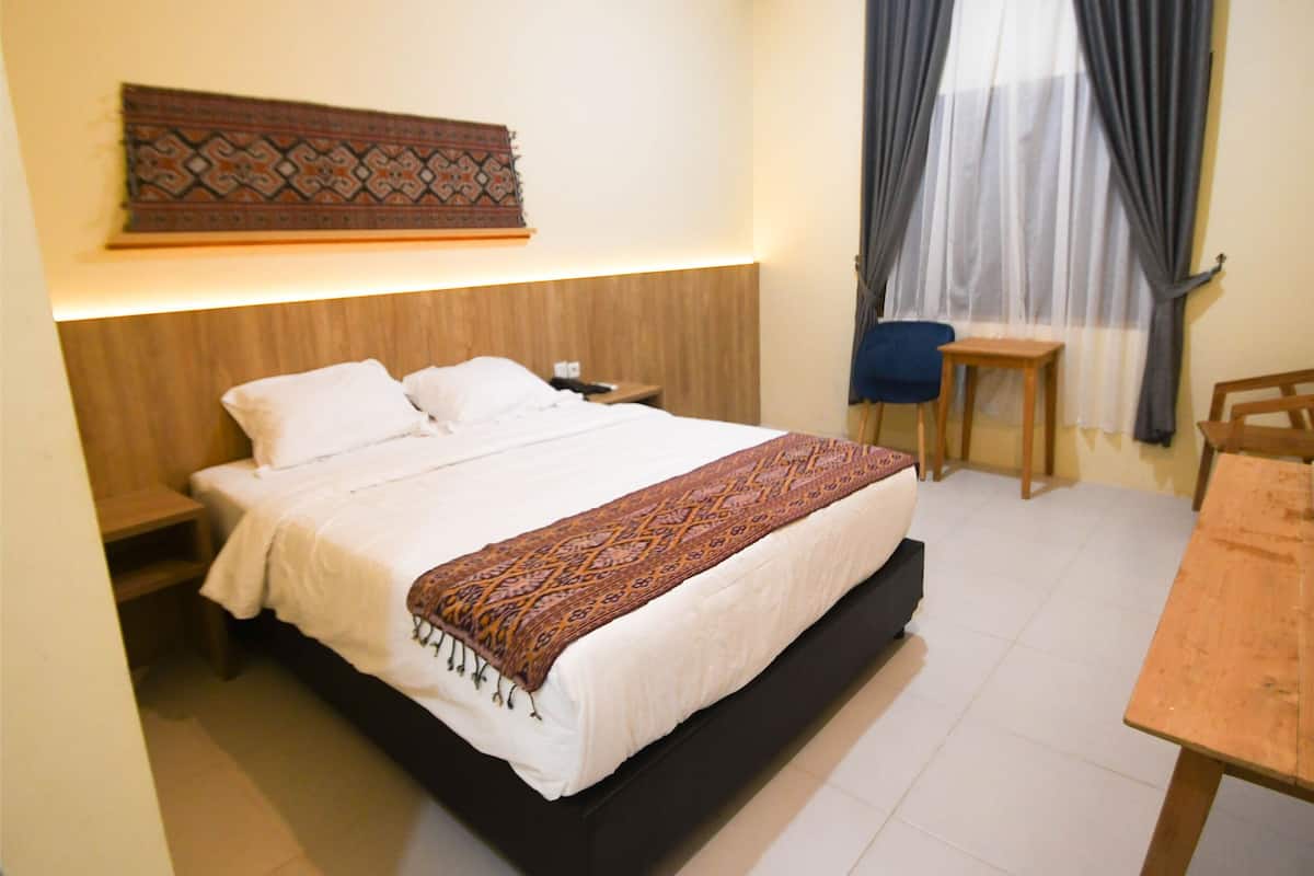 A neatly made bed with a patterned throw, a bedside table, a chair, and a wooden table in a room with a simple layout and a plain wall.