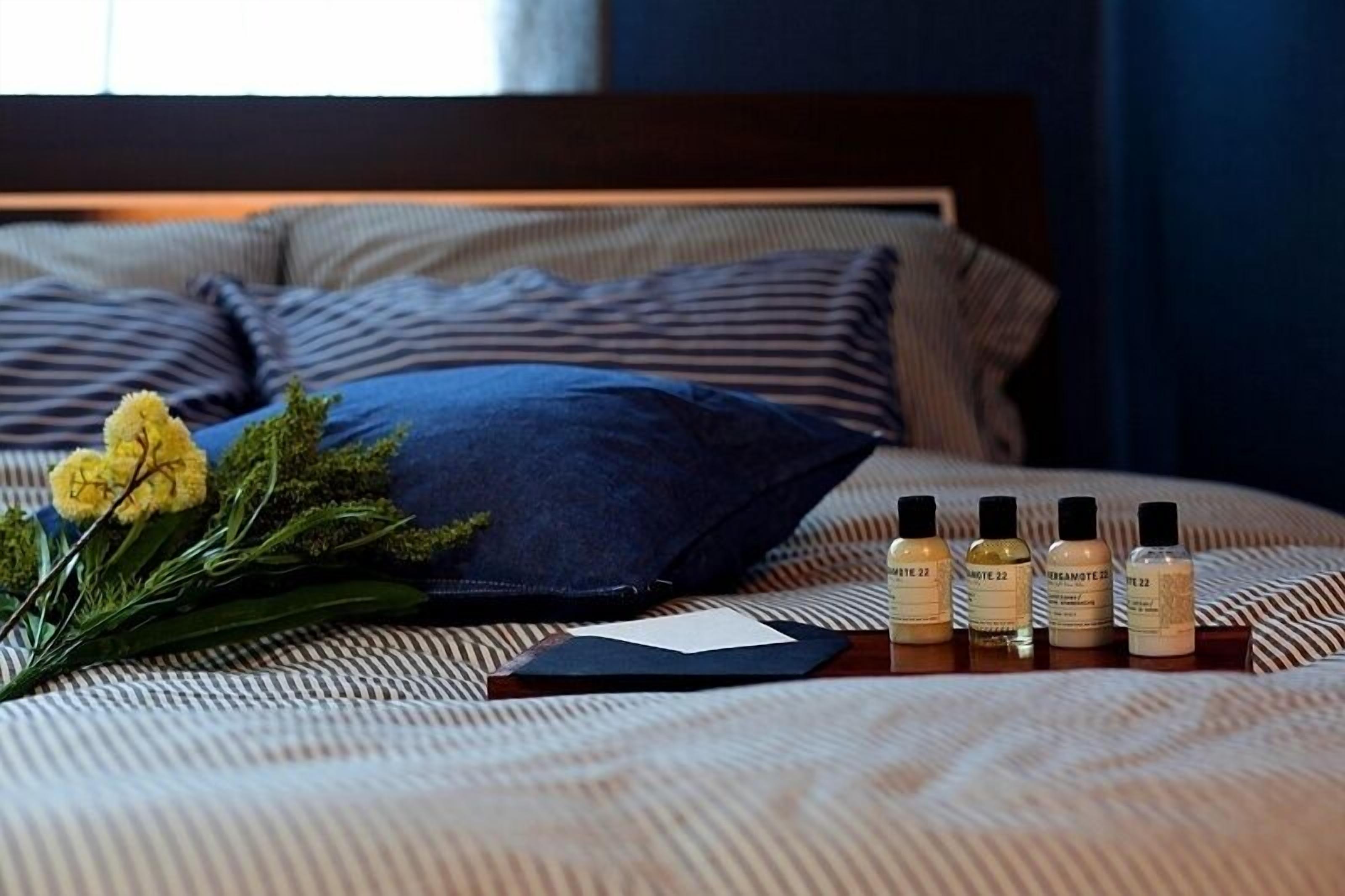 Cama bem arrumada com travesseiro listrado, um buquê de flores e três frascos de loção corporal em uma mesa de cabeceira.
