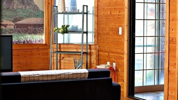 Ein gemütliches Zimmer mit Holzwänden, einem schwarzen Sofa, einem Metallregal mit Pflanzen und einem großen Fenster mit Blick auf Grünflächen.