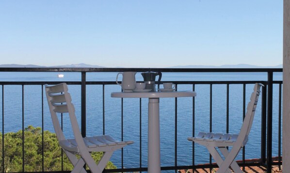 Un balcon avec deux chaises blanches et une petite table sur laquelle se trouve une cafetière, donnant sur une mer calme et des montagnes lointaines.