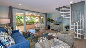 Ein modernes Wohnzimmer mit einem blauen Sofa, gemusterten Kissen, einem Couchtisch und einem Fernseher. Eine Wendeltreppe und ein Balkon mit Blick auf Grünpflanzen sind ebenfalls vorhanden.