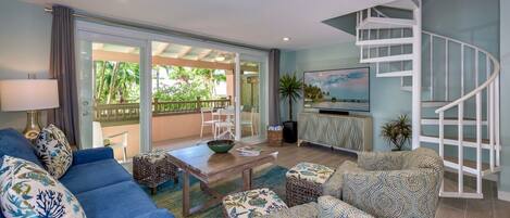 A modern living room with a blue sofa, patterned cushions, a coffee table, and a TV. There's a spiral staircase and a balcony with a view of greenery.