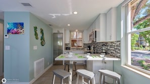 A modern kitchen with white cabinets, a stainless steel refrigerator, a dining area with stools, and a large window offering a view of the outside. - **DOCKSIDE DAYDREAMS @ COCONUT MALLORY** Villa + Last Key White Glove Service... (Key West)