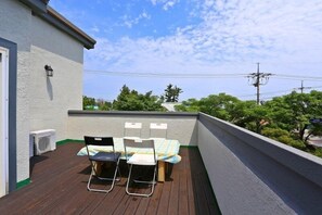 A balcony with a table and chairs, a white wall, and a view of trees and power lines. - Munmoon House (Seogwipo)