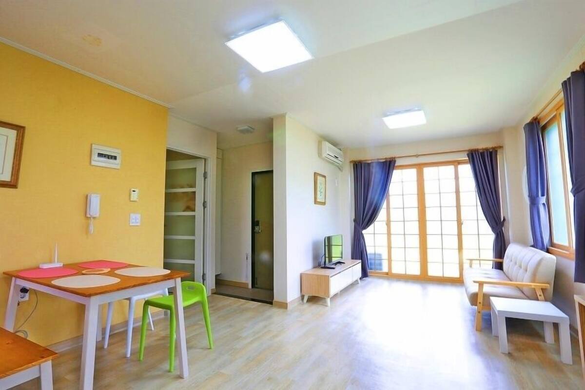 Ein helles Wohnzimmer mit einer Couch, einem kleinen Tisch und einem Fernseher. Eine Tür führt in einen anderen Raum, und ein Fenster ist mit Vorhängen drapiert.
