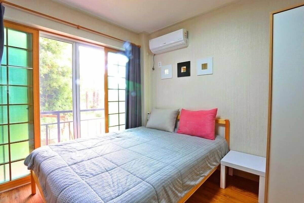Ein Schlafzimmer mit einem Bett, einem kleinen Nachttisch, einem Fenster mit Blick ins Grüne und einer Klimaanlage an der Wand.