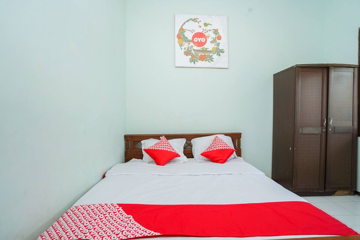A bedroom with a bed, two pillows, a red and white blanket, a wooden wardrobe, and a wall decoration.
