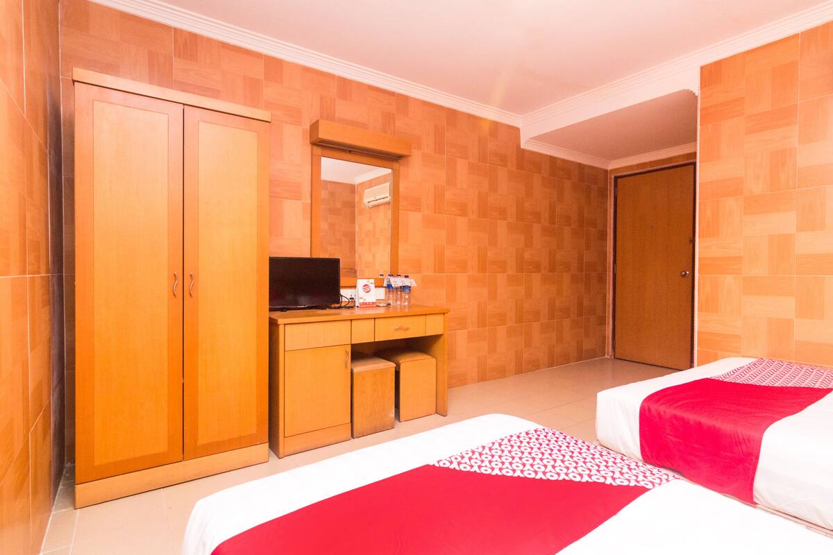 Een hotelkamer met een houten garderobe, een bureau met spiegel, een bed met rood-witte dekbedovertrek en een kamerbreed tapijt met dessin.