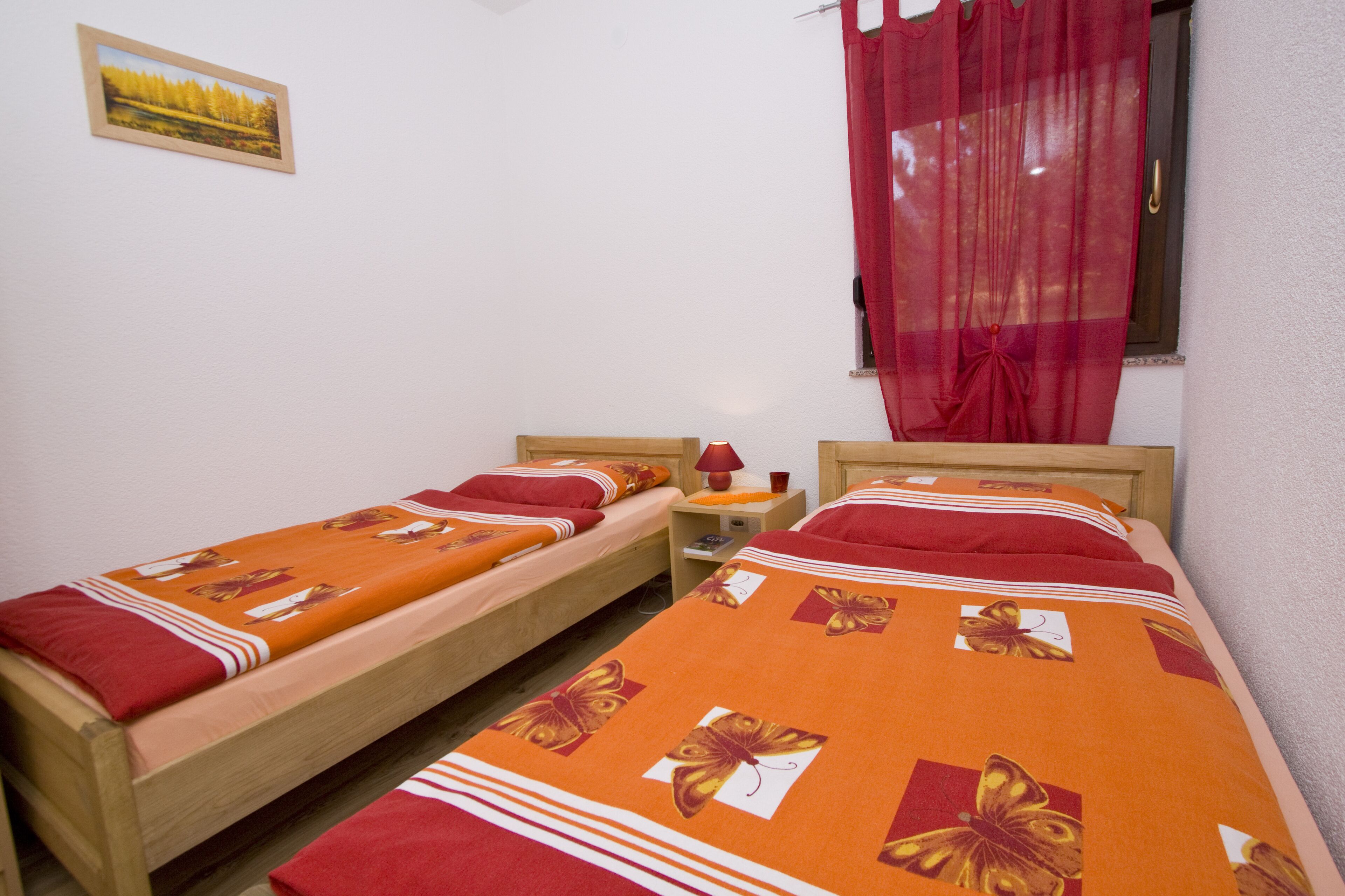 A room with two single beds, each with a red and orange floral bedspread, a small bedside table with a lamp, and a window with red curtains.