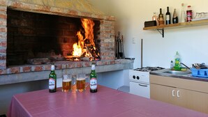 Une cheminée en pierre avec un feu, une table avec des bouteilles de bière et des verres, et un coin cuisine avec une cuisinière à gaz et des étagères remplies de bouteilles.
