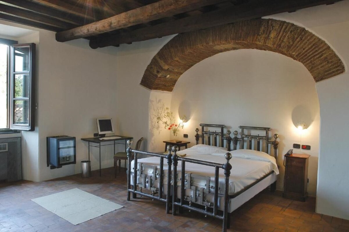 Une chambre avec une arche en pierre, un lit avec un cadre en métal, un bureau avec un ordinateur et une petite table avec une lampe.
