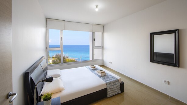A bedroom with a large window, a bed, a bedside table, a mirror, and a view of the sea. - 1 Bedroom apartment located in the heart of protaras. (Protaras)