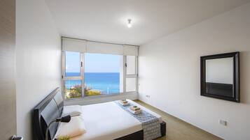 Un dormitorio con un ventanal grande, una cama, una mesita de noche, un espejo y vistas al mar.