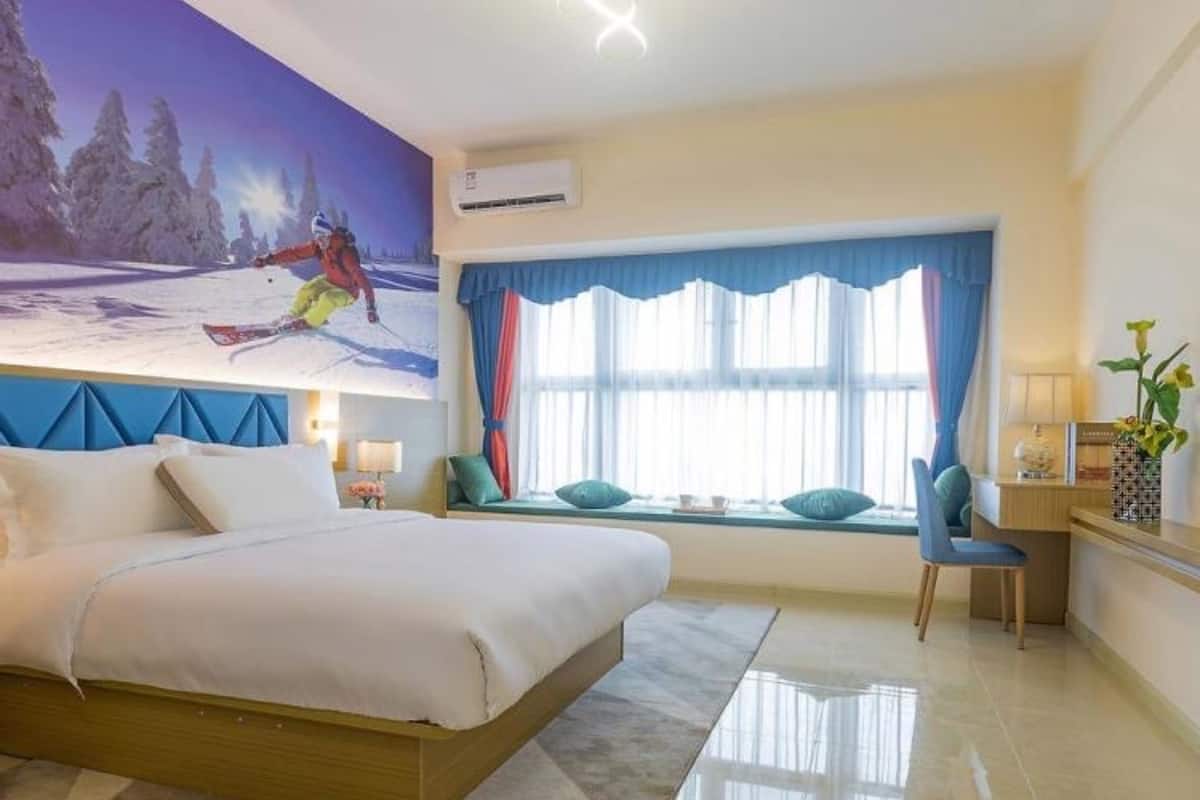 A hotel room with a large bed, a window with curtains, a desk with a chair, and a wall mural of a skier.