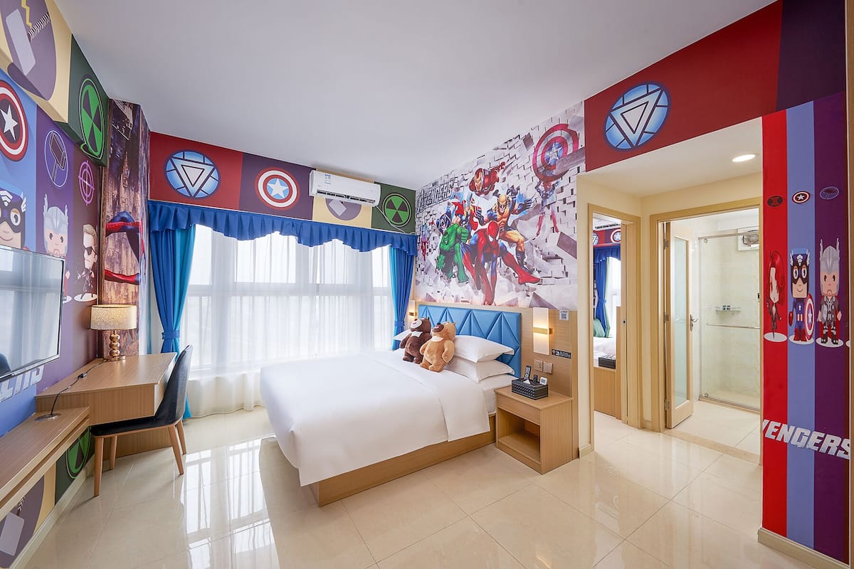A hotel room with a large bed, a desk, and a shower. The walls are decorated with superhero-themed artwork.