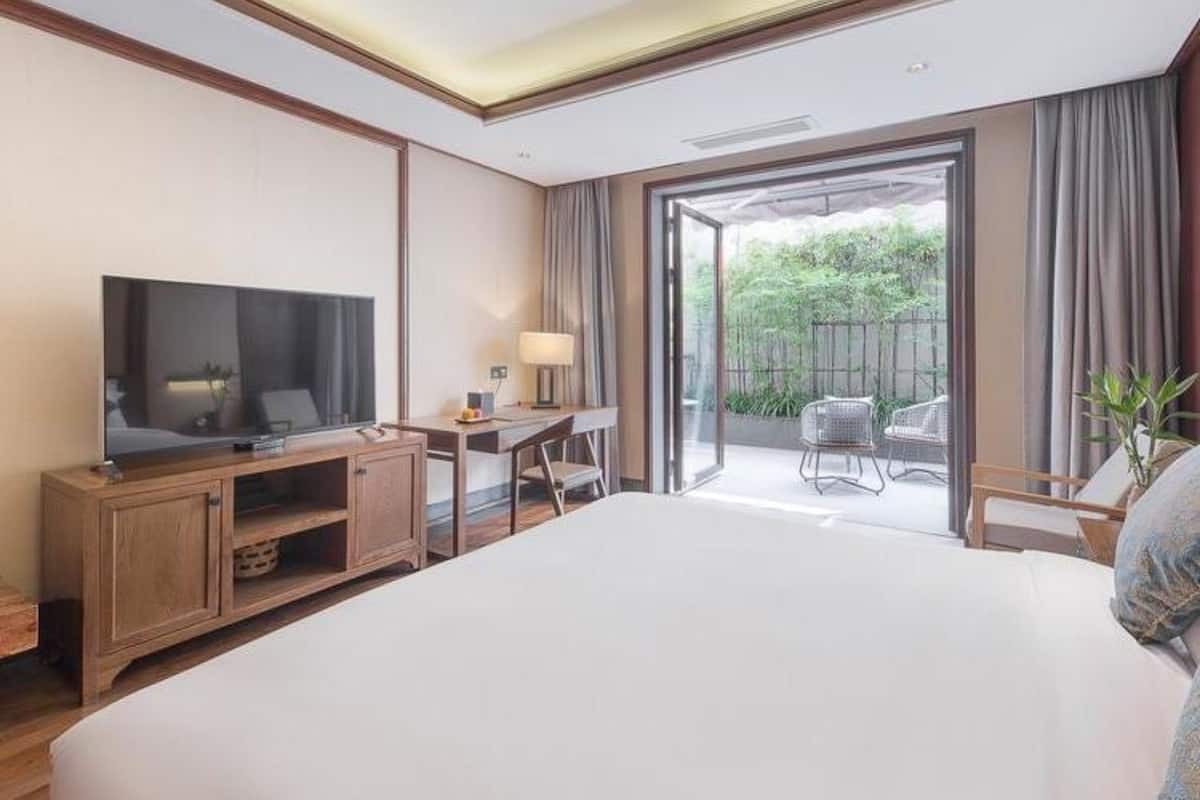 A modern hotel room with a large bed, a TV, a desk, and a balcony with outdoor seating.